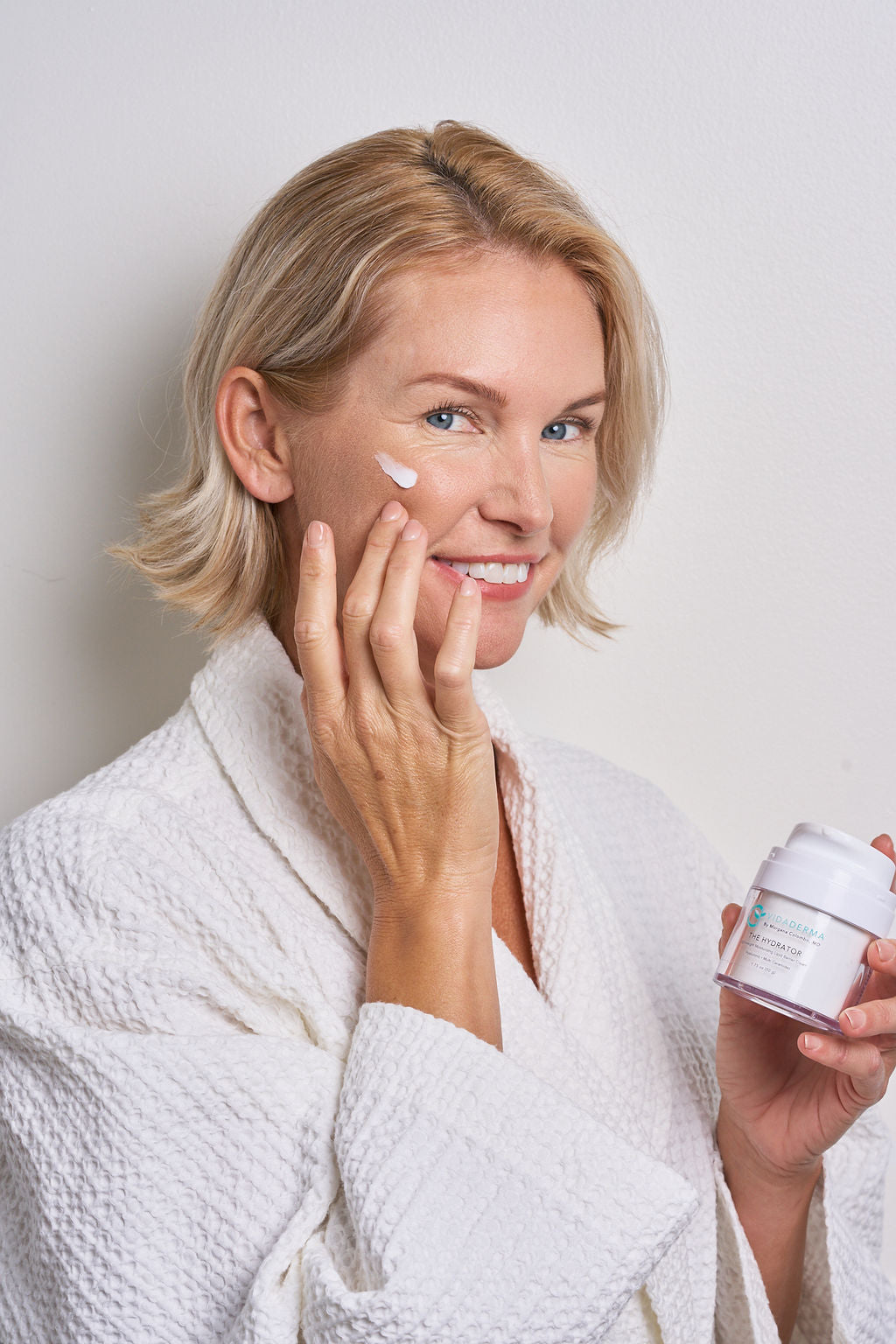 A smiling blonde woman in a white textured robe applies cream to her cheek while holding a small white cosmetic jar against a light gray background.