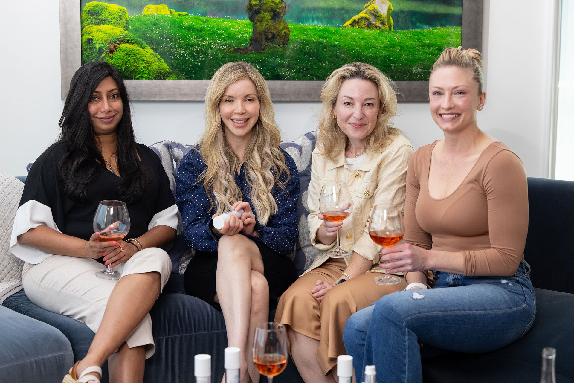 Four women sitting on a couch with wine glasses, smiling at the camera.