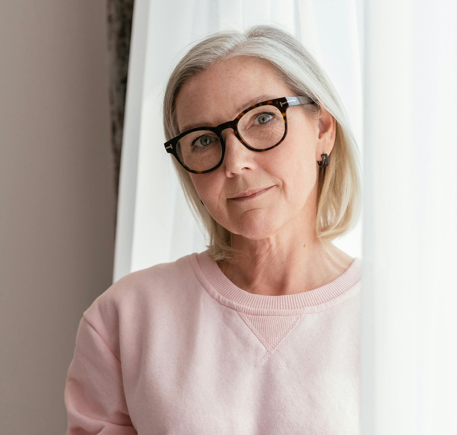 Woman wearing glasses and a pink sweater standing in front of a white curtain.