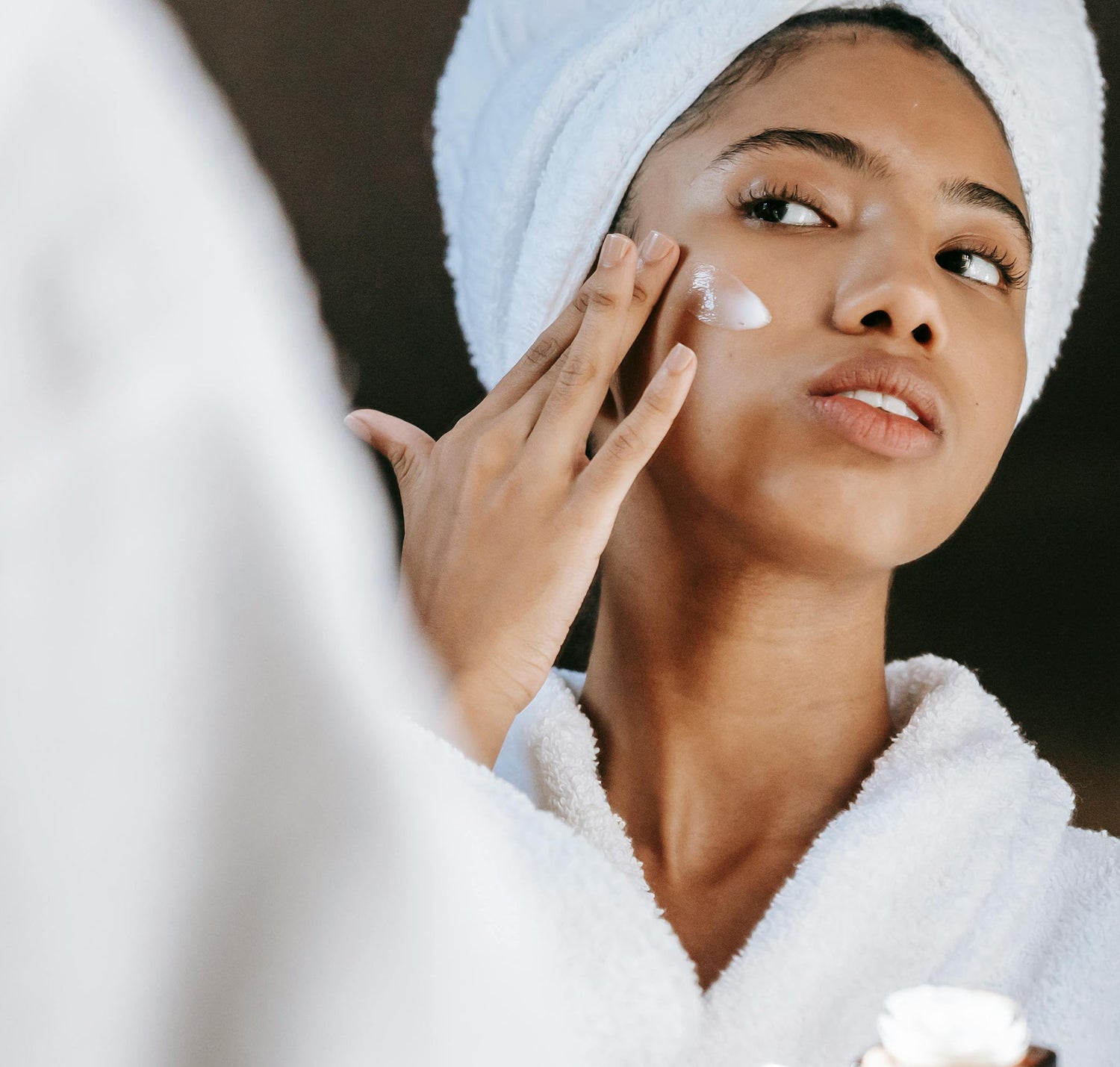 Woman applying cream to her face with a towel on her head, wearing a white robe.