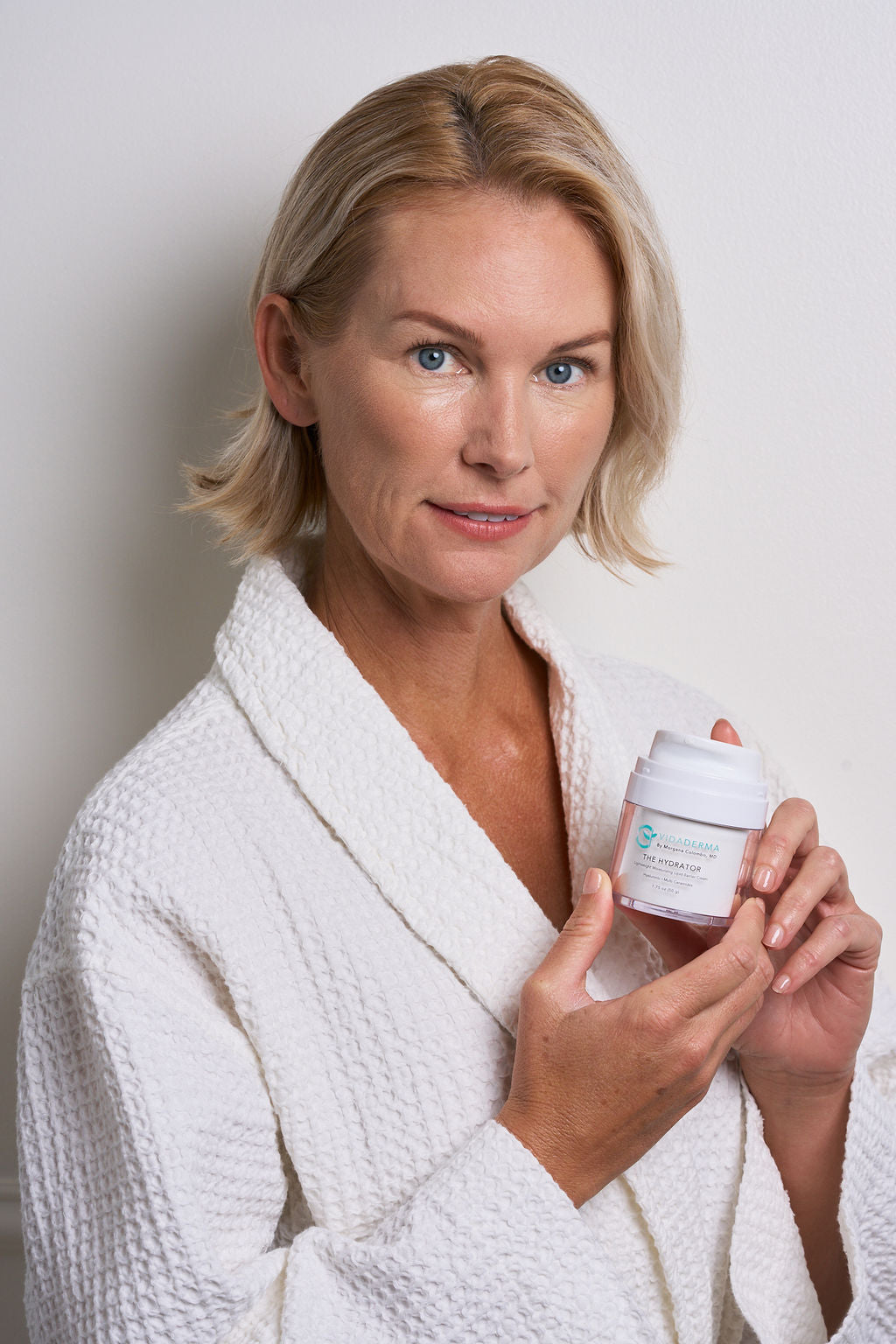 A blonde woman in a white textured bathrobe holds a small white cosmetic jar with a white lid against a plain light gray background.